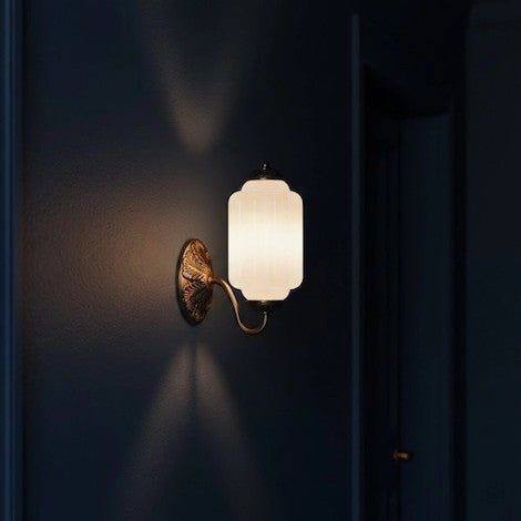 A vintage-inspired milk glass and oiled brass wall sconce glowing against a dark navy blue wall.